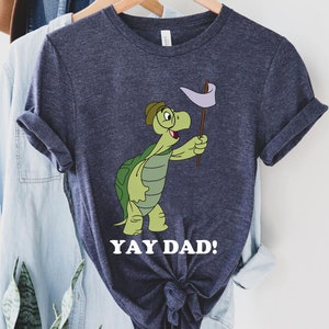 May include: Navy blue t-shirt featuring a cartoon turtle holding a flag. The turtle is green with a brown hat and is smiling. The text "YAY DAD!" is printed in white below the turtle.