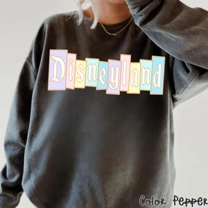 Puede incluir: Una sudadera gris oscuro con la palabra "Disneyland" en colores pastel en el pecho. El texto est&aacute; en un estilo de letras en bloque con un contorno dorado. La esquina inferior derecha de la sudadera tiene el texto "Color: Pepper".