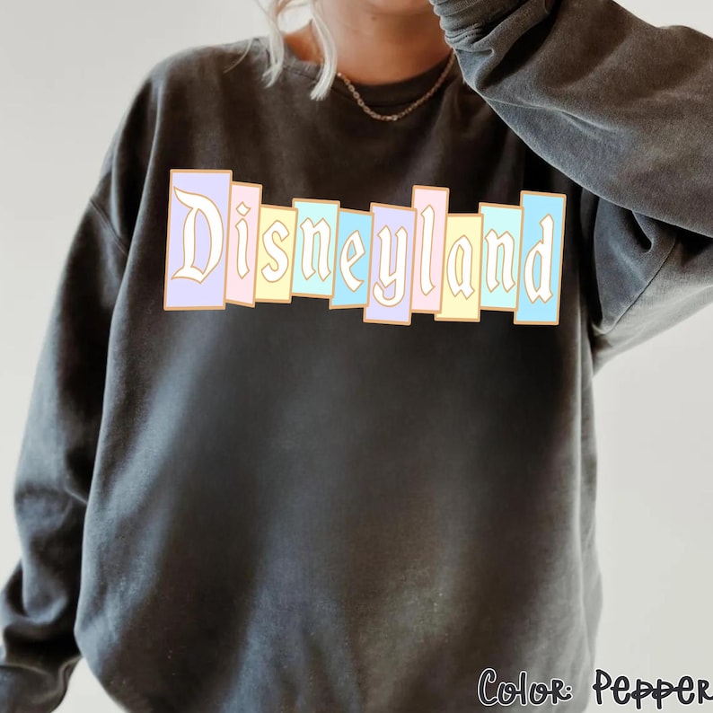 Puede incluir: Una sudadera gris oscuro con la palabra "Disneyland" en colores pastel en el pecho. El texto est&aacute; en un estilo de letras en bloque con un contorno dorado. La esquina inferior derecha de la sudadera tiene el texto "Color: Pepper".