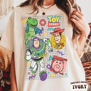 May include: Ivory-colored t-shirt featuring a colorful graphic of Toy Story characters, including Buzz Lightyear, Woody, Rex, and Hamm. The design is set against a grid background with geometric shapes. The words "TOY STORY" are displayed at the top.