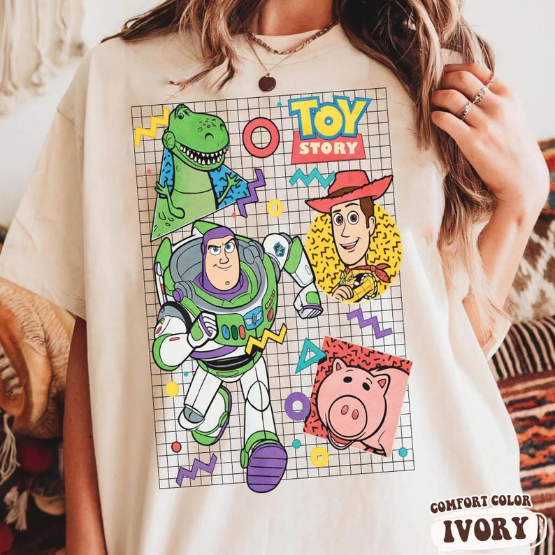 May include: Ivory-colored t-shirt featuring a colorful graphic of Toy Story characters, including Buzz Lightyear, Woody, Rex, and Hamm. The design is set against a grid background with geometric shapes. The words "TOY STORY" are displayed at the top.