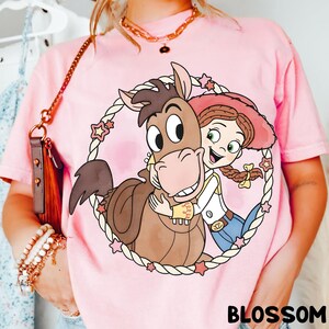 May include: Pink t-shirt featuring a cartoon graphic of a horse and a cowgirl embracing within a rope circle with star accents. The word "BLOSSOM" is printed in black at the bottom of the shirt.