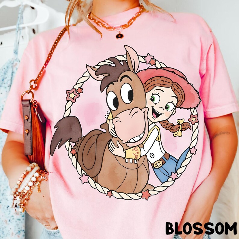 May include: Pink t-shirt featuring a cartoon graphic of a horse and a cowgirl embracing within a rope circle with star accents. The word "BLOSSOM" is printed in black at the bottom of the shirt.