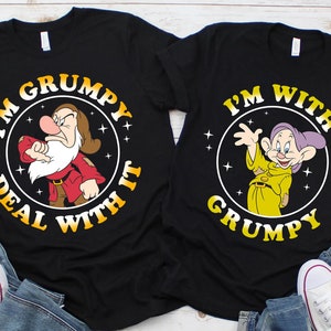 Op de afbeelding: Twee zwarte T-shirts met een grafische print van personages uit de film Sneeuwwitje. Het shirt aan de linkerkant zegt "I'm Grumpy Deal With It" en toont Grumpy. Het shirt aan de rechterkant zegt "I'm With Grumpy" en toont Dopey.