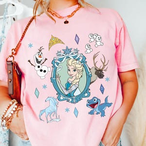 Puede incluir: Camiseta rosa con un estampado gr&aacute;fico de una imagen de dibujos animados de Elsa de Frozen. El gr&aacute;fico tambi&eacute;n incluye a Olaf, Sven el reno y una salamandra azul.