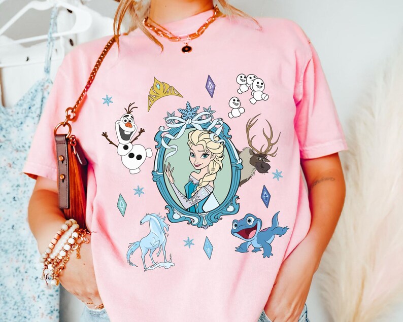 Puede incluir: Camiseta rosa con un estampado gr&aacute;fico de una imagen de dibujos animados de Elsa de Frozen. El gr&aacute;fico tambi&eacute;n incluye a Olaf, Sven el reno y una salamandra azul.