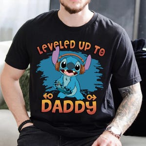 Puede incluir: Camiseta negra con un personaje azul Stitch que lleva auriculares y sostiene un mando de videojuego. El texto "Leveled Up To Daddy" est&aacute; impreso en la camiseta.