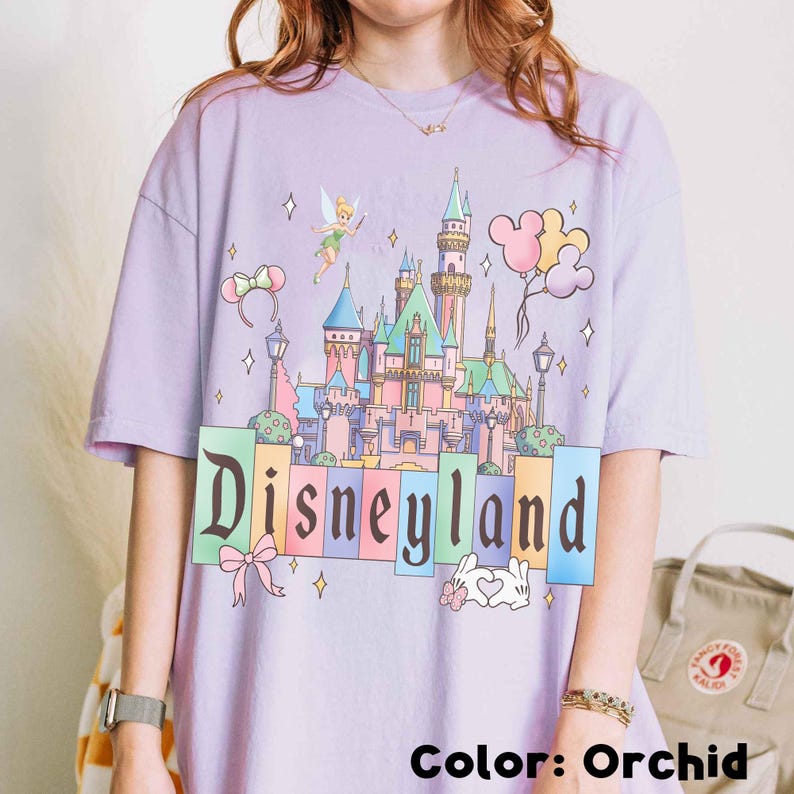 Puede incluir: Camiseta color orqu&iacute;dea con un gr&aacute;fico del castillo de Disneyland. El dise&ntilde;o incluye a Campanilla, globos de Mickey Mouse y la palabra "Disneyland". La camiseta tiene un corte holgado.