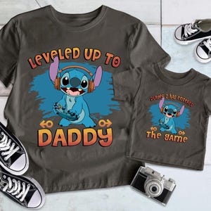Puede incluir: Dos camisetas grises con un personaje azul Stitch de Lilo & Stitch. La camiseta m&aacute;s grande dice "Leveled Up To Daddy" y la m&aacute;s peque&ntilde;a dice "Player 2 Has Entered The Game".