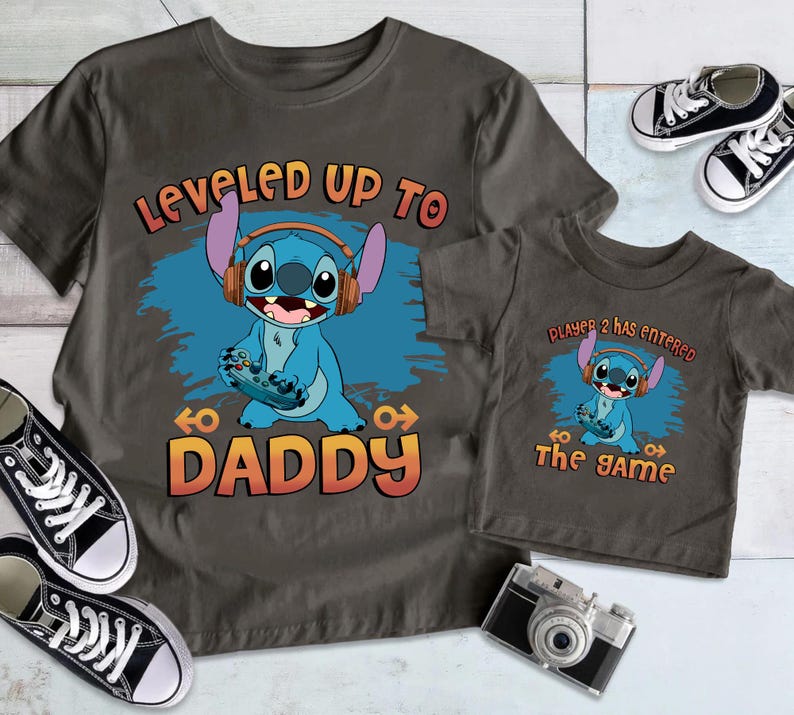 Puede incluir: Dos camisetas grises con un personaje azul Stitch de Lilo & Stitch. La camiseta m&aacute;s grande dice "Leveled Up To Daddy" y la m&aacute;s peque&ntilde;a dice "Player 2 Has Entered The Game".