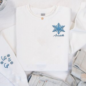 May include: White sweatshirt with blue snowflake embroidery and the text "Let it Go" on the sleeves. The snowflake is on the front of the shirt with the text "Arendelle" underneath.