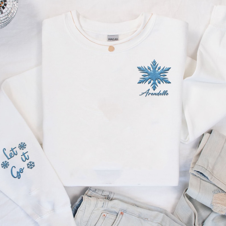May include: White sweatshirt with blue snowflake embroidery and the text "Let it Go" on the sleeves. The snowflake is on the front of the shirt with the text "Arendelle" underneath.