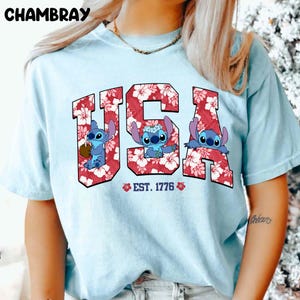 May include: Light blue t-shirt featuring the word "USA" in red floral print with three cartoon Stitch characters. The text "EST. 1776" is below the word "USA". The word "CHAMBRAY" is at the top left.