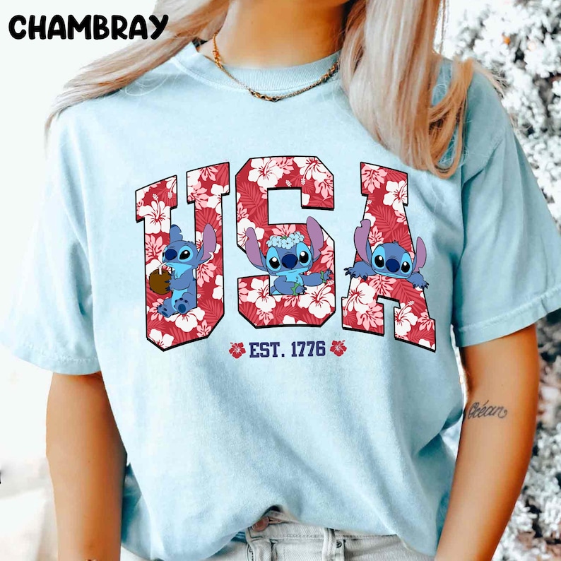 May include: Light blue t-shirt featuring the word "USA" in red floral print with three cartoon Stitch characters. The text "EST. 1776" is below the word "USA". The word "CHAMBRAY" is at the top left.
