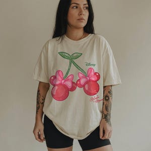 Op de afbeelding: Cr&egrave;mekleurig T-shirt met een cartoon kers ontwerp met Minnie Mouse oren en strikken. De kersen zijn rood met roze strikken en groene stelen. Het Disney-logo staat boven de kersen en "Minnie Mouse" staat eronder.