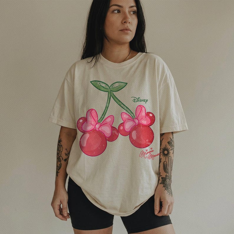 Op de afbeelding: Cr&egrave;mekleurig T-shirt met een cartoon kers ontwerp met Minnie Mouse oren en strikken. De kersen zijn rood met roze strikken en groene stelen. Het Disney-logo staat boven de kersen en "Minnie Mouse" staat eronder.
