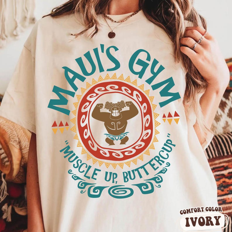 May include: Ivory-colored t-shirt with the text "MAUI'S GYM" in teal. A circular graphic features a cartoon character flexing, surrounded by a red and yellow design. The phrase "MUSCLE UP, BUTTERCUP" is below the graphic. The shirt is labeled "COMFORT COLOR IVORY".