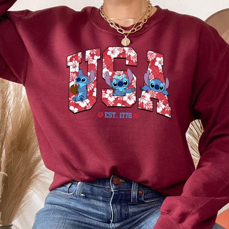 May include: Burgundy sweatshirt featuring the word "USA" in a floral pattern with three cartoon characters. The sweatshirt includes the text "EST. 1776" below the letters. The design is centered on the front of the garment.