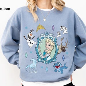 Puede incluir: Una sudadera azul claro con un estampado gr&aacute;fico de personajes de la pel&iacute;cula Frozen. El estampado presenta a Elsa, Olaf y un reno. El texto "Blue Jean" est&aacute; impreso en la esquina superior izquierda.