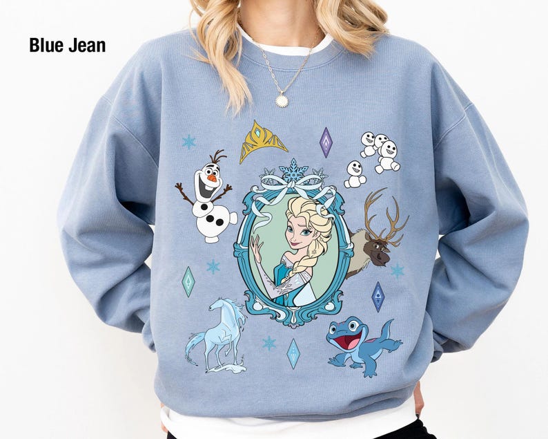 Puede incluir: Una sudadera azul claro con un estampado gr&aacute;fico de personajes de la pel&iacute;cula Frozen. El estampado presenta a Elsa, Olaf y un reno. El texto "Blue Jean" est&aacute; impreso en la esquina superior izquierda.