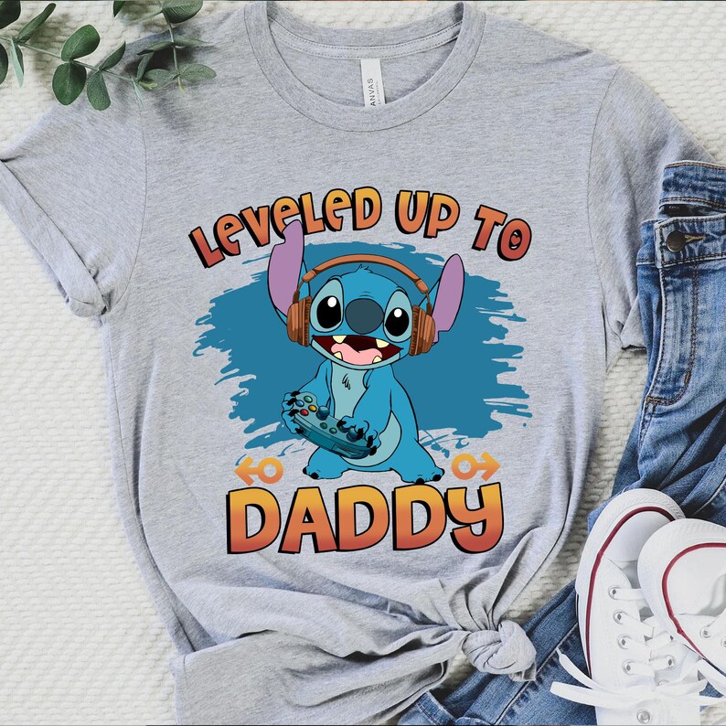 Puede incluir: Camiseta gris con un personaje azul Stitch con auriculares y sosteniendo un mando de videojuego. El texto "Leveled Up To Daddy" est&aacute; impreso en la camiseta.
