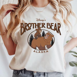 Puede incluir: Una camiseta blanca con un oso marrón y un cachorro de oso en su espalda. El texto "Disney's Brother Bear Alaska" está impreso en la camiseta.