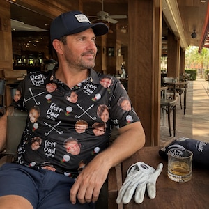 May include: A black golf shirt with the text "Best Dad by Par" and faces of children printed on it. The shirt also features golf balls and crossed golf clubs. A navy blue baseball cap with "MUN KIDS" is worn. A golf glove and a glass of amber liquid are on the table.