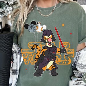 May include: A green t-shirt with a graphic of Tigger dressed as Darth Vader from Star Wars. Tigger is holding a lightsaber and has a Death Star behind him. The text "Star Wars" is printed in orange on the shirt.