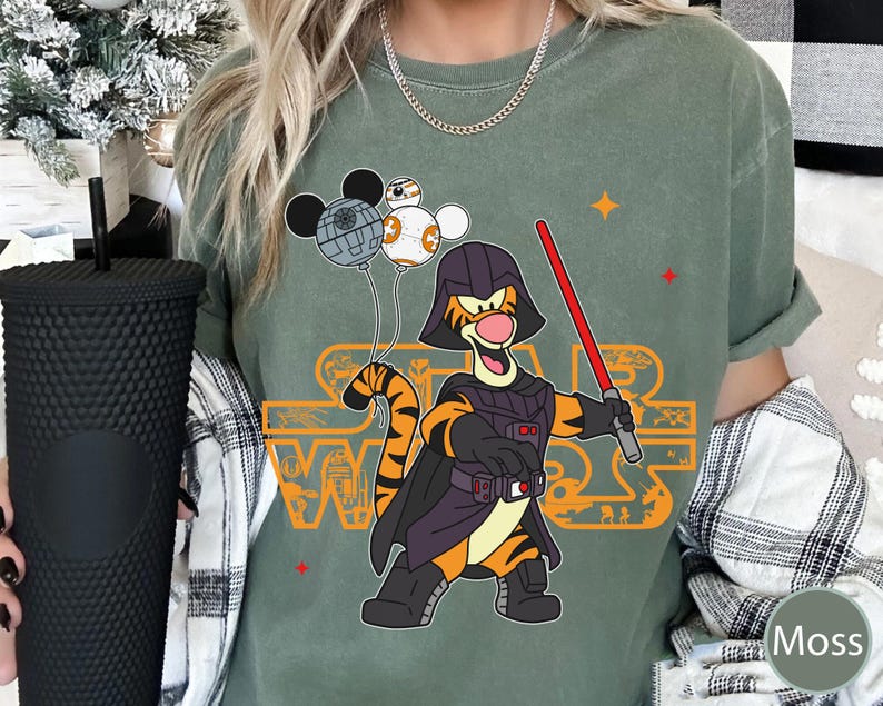 May include: A green t-shirt with a graphic of Tigger dressed as Darth Vader from Star Wars. Tigger is holding a lightsaber and has a Death Star behind him. The text "Star Wars" is printed in orange on the shirt.