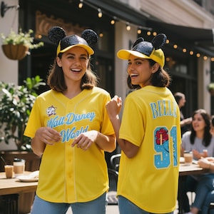 May include: Two people wearing yellow baseball jerseys with "Walt Disney World" and "Bella 91" on the back. They are also wearing yellow caps with black sequined mouse ears. The jerseys have a white trim.