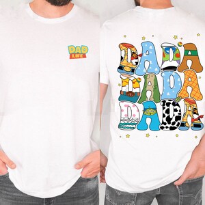 Op de afbeelding: Wit T-shirt met de tekst "DAD LIFE" op de voorkant en "DADA DADA DADA" op de achterkant. De tekst op de achterkant is in een kleurrijk, cartoonachtig lettertype met steraccenten. Een casual kledingstuk voor dagelijks gebruik.