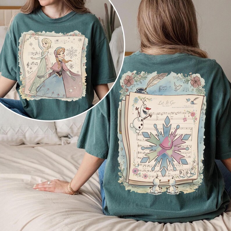 May include: Teal-colored t-shirt featuring a colorful illustration of characters from a popular animated film. The front design shows two characters with musical notes and snowflakes. The back design includes sheet music, a snowman, and the words "Let It Go".