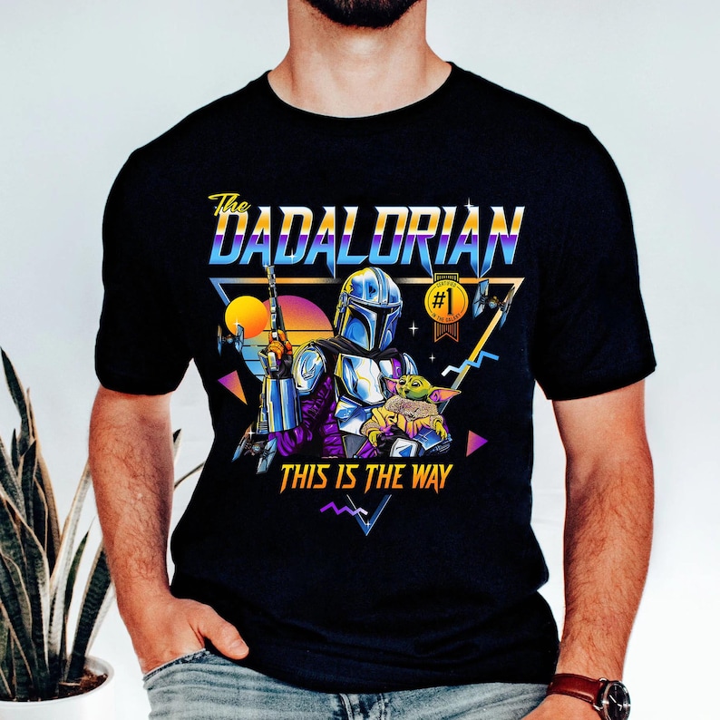 May include: A black t-shirt with a retro-style graphic of a Mandalorian character holding a small green creature. The graphic includes the text "The Dadalorian" and "This is the Way".