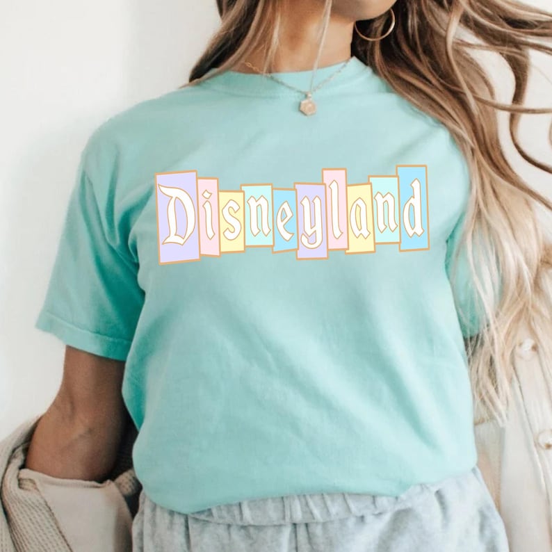Puede incluir: Una camiseta azul claro con la palabra "Disneyland" impresa en el pecho en un dise&ntilde;o de bloques de color pastel. El texto est&aacute; en una fuente vintage y la camiseta tiene mangas cortas.