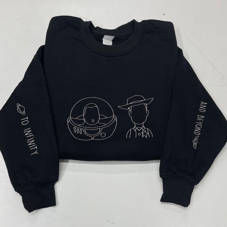 May include: Black sweatshirt with white embroidered outlines of Buzz Lightyear and Sheriff Woody. The left sleeve has the text "TO INFINITY" and a planet. The right sleeve has "AND BEYOND" and a rocket.