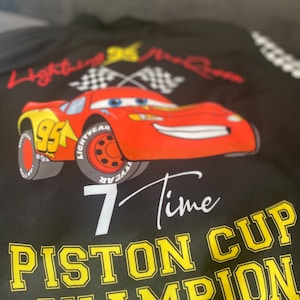 May include: Black t-shirt with a red race car graphic, checkered flags, and the text "Lightning McQueen 7 Time Piston Cup Champion".