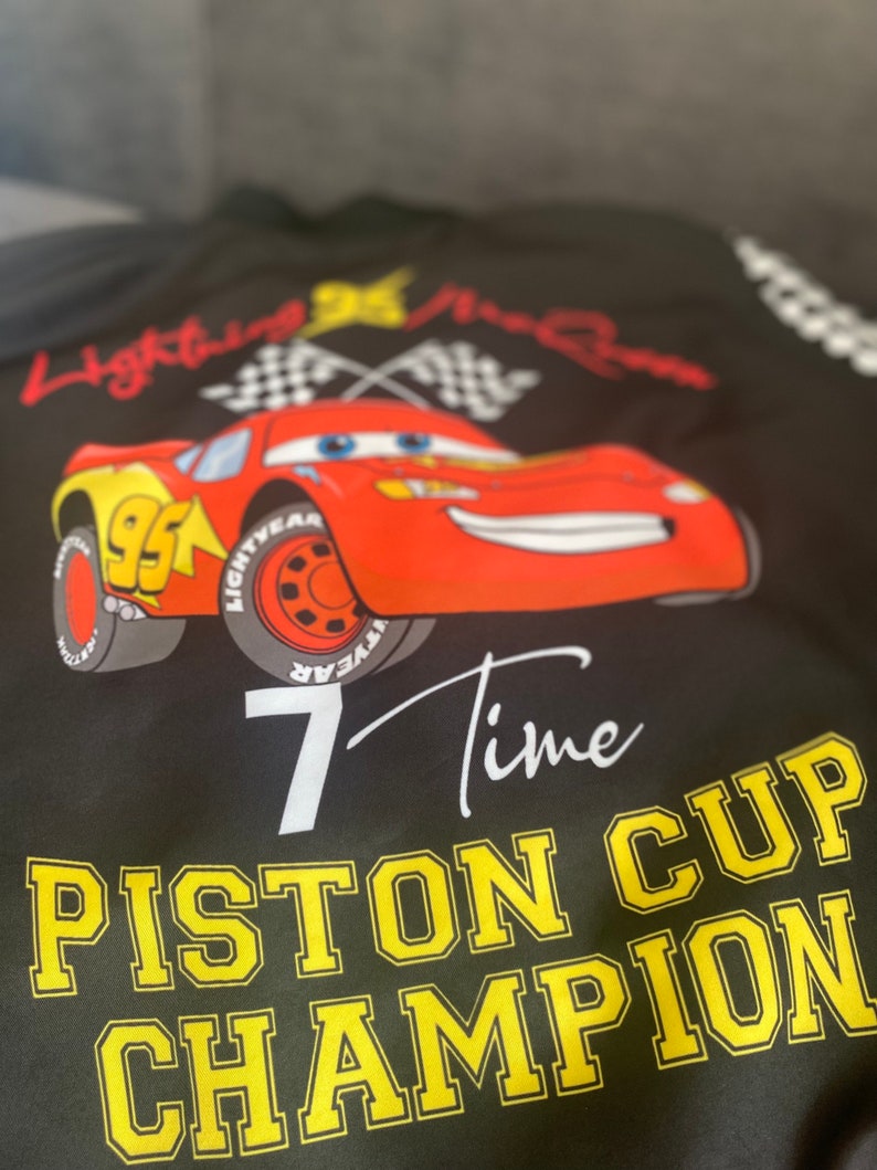 May include: Black t-shirt with a red race car graphic, checkered flags, and the text "Lightning McQueen 7 Time Piston Cup Champion".