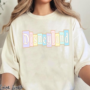 Puede incluir: Camiseta color marfil con la palabra "Disneyland" en colores pastel. El texto, en una fuente vintage, est&aacute; segmentado en rect&aacute;ngulos verticales rosas, amarillos y azules. La camiseta tiene un corte holgado.