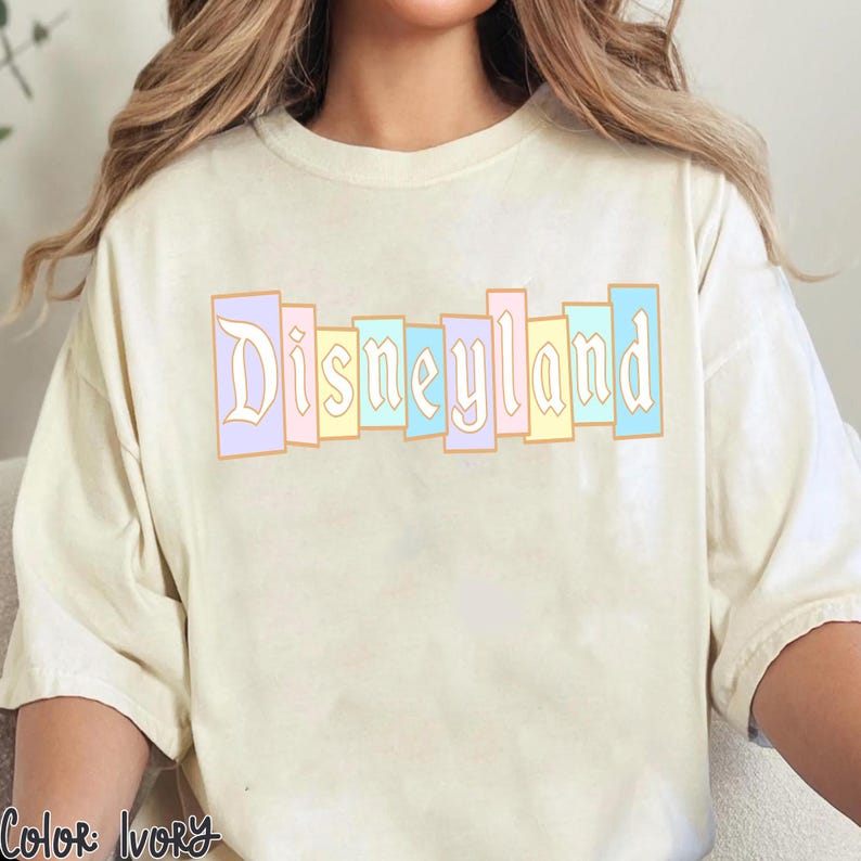 Puede incluir: Camiseta color marfil con la palabra "Disneyland" en colores pastel. El texto, en una fuente vintage, est&aacute; segmentado en rect&aacute;ngulos verticales rosas, amarillos y azules. La camiseta tiene un corte holgado.
