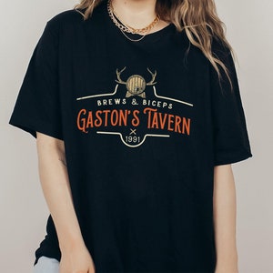 Op de afbeelding: Zwart T-shirt met een vintage-stijl logo met de tekst "Brews & Biceps Gaston's Tavern 1991" met een afbeelding van een hertenkop en een vat.
