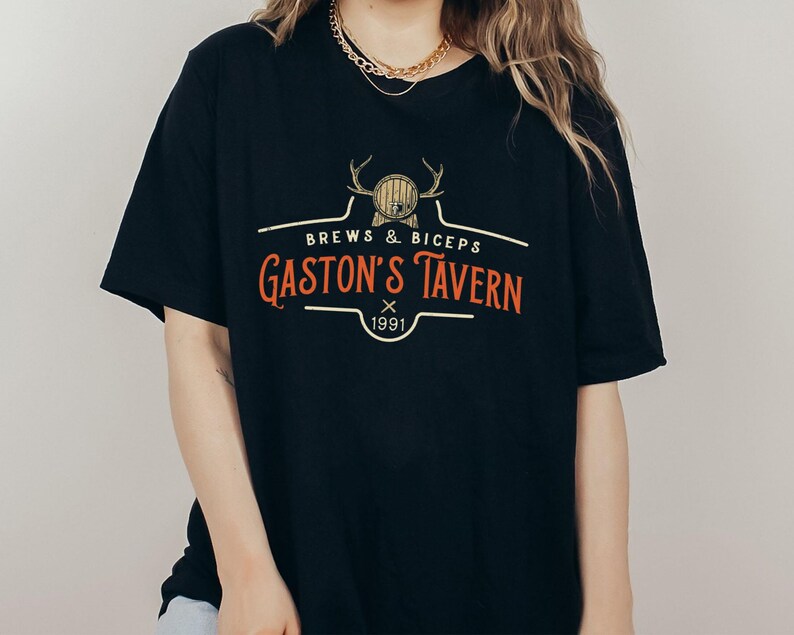 Op de afbeelding: Zwart T-shirt met een vintage-stijl logo met de tekst "Brews & Biceps Gaston's Tavern 1991" met een afbeelding van een hertenkop en een vat.