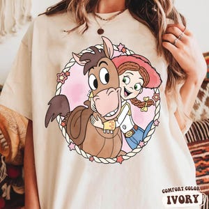 May include: Ivory-colored t-shirt featuring a cartoon illustration of a cowgirl hugging a horse inside a pink heart-shaped frame with stars. The shirt has the words "Comfort Color Ivory" printed at the bottom.