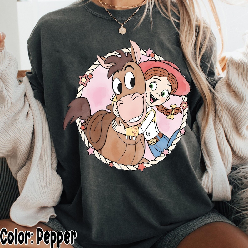 May include: Pepper-colored sweatshirt featuring a circular graphic of a cartoon horse hugging a cowgirl. The graphic is surrounded by a rope-like border with pink stars. The text "Color: Pepper" is at the bottom of the image.