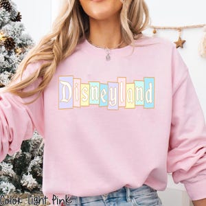 Puede incluir: Sudadera rosa claro con la palabra "Disneyland" en colores pastel. El texto est&aacute; en una fuente vintage y est&aacute; delineado en dorado. La sudadera es de cuello redondo y tiene mangas largas.