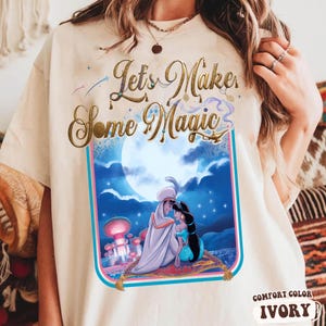 May include: Ivory-colored t-shirt featuring a graphic of Aladdin and Jasmine on a magic carpet, with a full moon backdrop. The text "Let's Make Some Magic" is displayed in gold script above the image. The shirt is labeled "Comfort Color Ivory".
