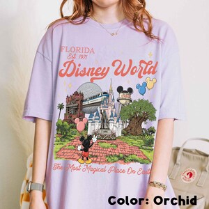 Puede incluir: Camiseta color orqu&iacute;dea con un gr&aacute;fico colorido de Disney World. El dise&ntilde;o incluye el texto "FLORIDA Est. 1971" y "Disney World", junto con ilustraciones de lugares emblem&aacute;ticos del parque y Mickey Mouse. Tambi&eacute;n incluye la frase "The Most Magical Place On Earth".
