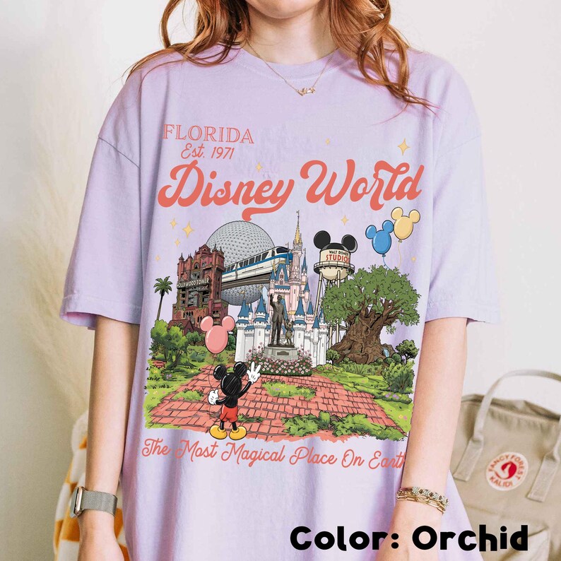 Puede incluir: Camiseta color orqu&iacute;dea con un gr&aacute;fico colorido de Disney World. El dise&ntilde;o incluye el texto "FLORIDA Est. 1971" y "Disney World", junto con ilustraciones de lugares emblem&aacute;ticos del parque y Mickey Mouse. Tambi&eacute;n incluye la frase "The Most Magical Place On Earth".