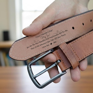 Puede incluir: Un cinturón de cuero marrón con hebilla plateada. El interior del cinturón está grabado con un mensaje de San Valentín, incluyendo las palabras "Happy Valentine's Day!" y "I Love You Aria."