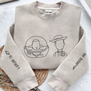 May include: Beige sweatshirt featuring black line art of Buzz Lightyear in his space suit and Sheriff Woody. The left sleeve says "TO INFINITY" and the right sleeve says "AND BEYOND". A fun, casual top.
