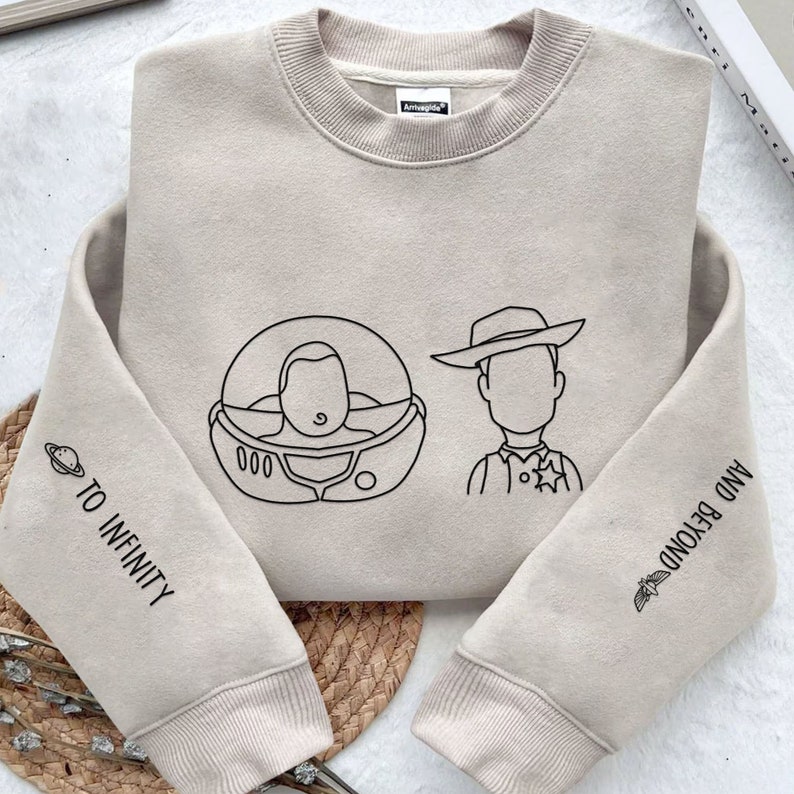May include: Beige sweatshirt featuring black line art of Buzz Lightyear in his space suit and Sheriff Woody. The left sleeve says "TO INFINITY" and the right sleeve says "AND BEYOND". A fun, casual top.